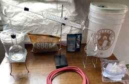 Photo of items arrayed on a wooden table, from right to left: a white five gallon bucket with the Williams Brewing logo on it and in front of that a plastic bag with the various bits of plumbing for it (it's a simple fermentation kit); a packet of Wyeast brand liquid yeast (for Saké & Spirits, so it can tolerate higher alcohol levels); a school-style distillation kit consisting of a Erlenmeyer flask on a stand, a Graham condenser (a large glass tube with a smaller helical glass tube inside it to condense vapor), a Kjeldahl distillation ball, rubber tube, and a spherical flat-bottomed boiling flask on a burner stand; a bag of brown sugar; and a smaller white bucket (around one gallon) containing five pounds of 3 Ångström synthetic zeolite molecular sieve pellets (for drying the alcohol.)